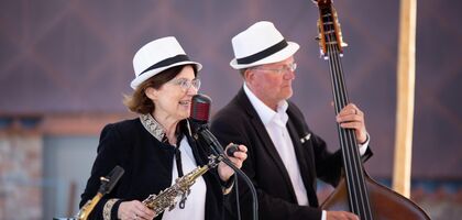 Eine Sängerin mit Klarinette in der Hand singt in eine fünfziger Jahre Mikrophone. rechts neben Ihr ein Mann mitt Kontrabass. Beide haben weiße Hüte auf dem Kopf, ein weißes Hemd an und schwarze Blaser