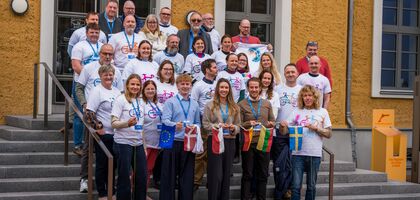 Eine Menschengruppe mit weißen T-shirts steht vor dem eingang eines Hauses (Vineta Bürgerhaus). die erste Reihe hält eine Kette aus kleinen Flaggen.