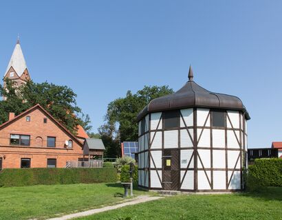 Achteckiges Häuschen Fachwerk weiß vermauert, im Hintergrund die Kirche