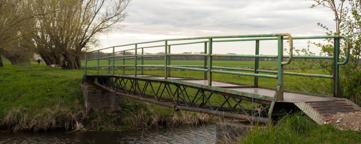 kleine schmale Eisen-Brücke über einen kleinen Fluss. Hinten links Kopfweiden. Umgebung mit Feldwiesen